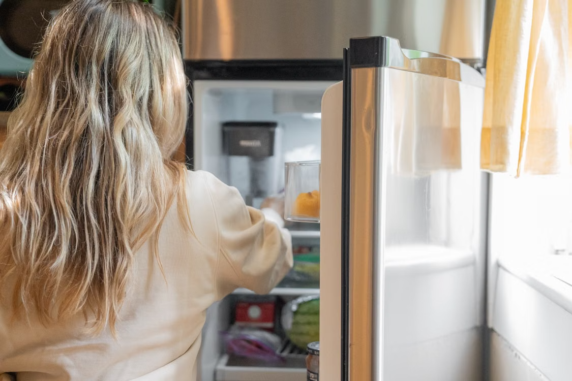 Back view of a blonde woman retrieving food from a refrigerator in a bright kitchen