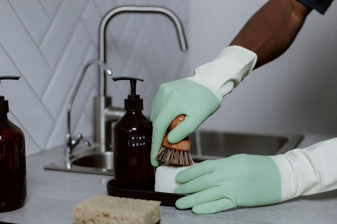 Hands in latex gloves washing dishes with soap at a kitchen sink.