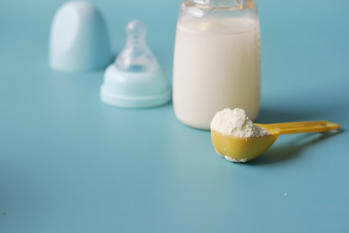 Close-up of baby milk formula powder in a yellow scoop with a bottle on a blue background